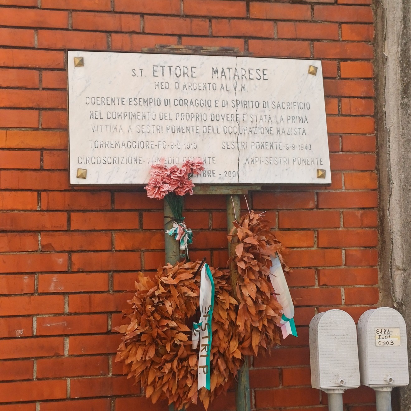 Lapide marmorea con iscrizione su parete di mattoni, sotto corone di fiori secchi appese.