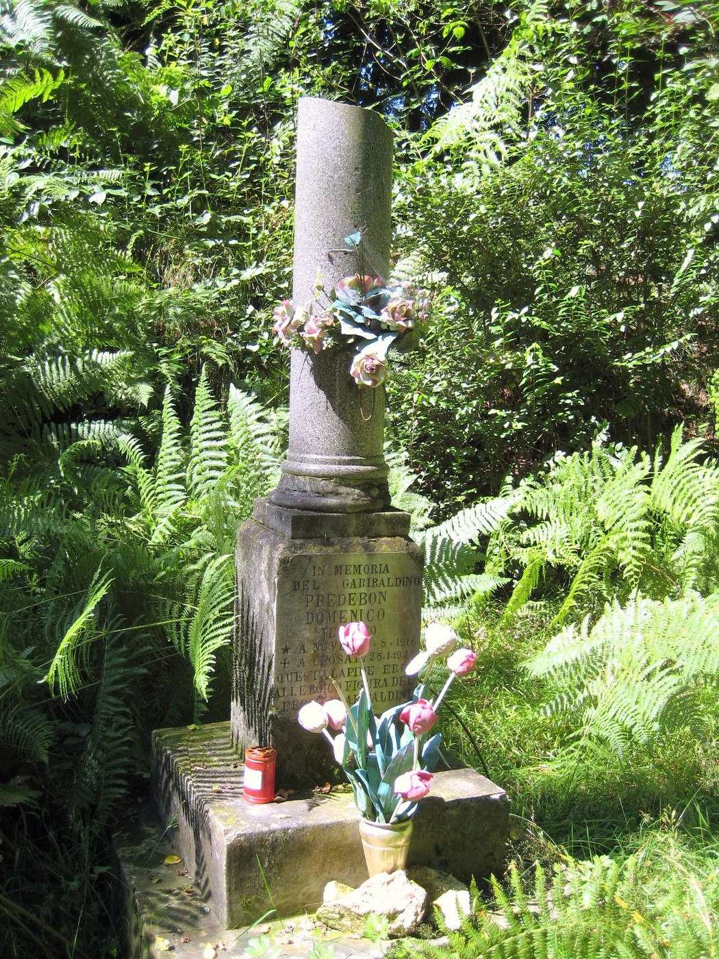 Cippo a Predebon, monumento circondato da vegetazione, adornato di fiori e candele.