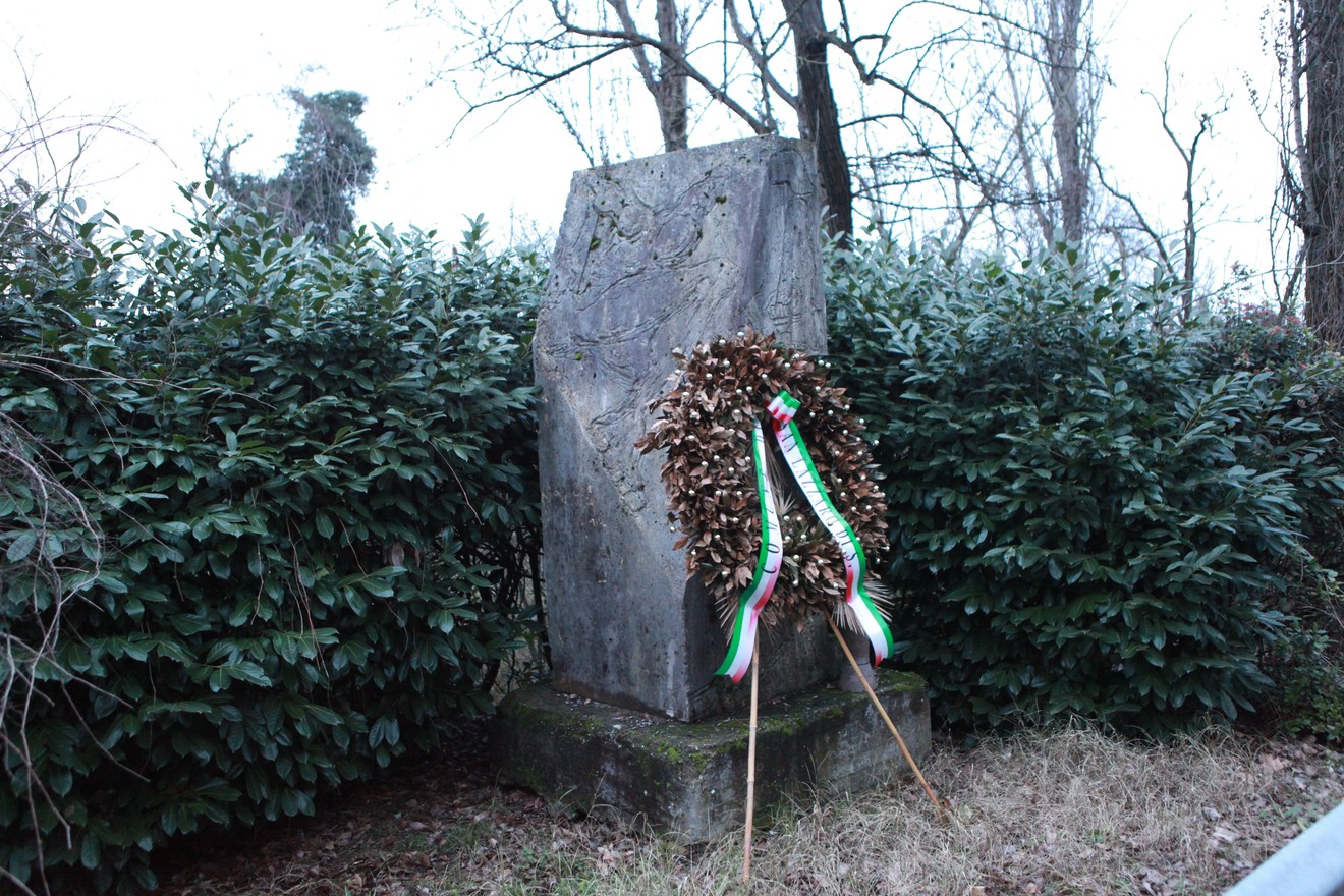 Il Monumento ai martiri di Pizzocalvo, con una corona di fiori, si staglia tra la vegetazione.