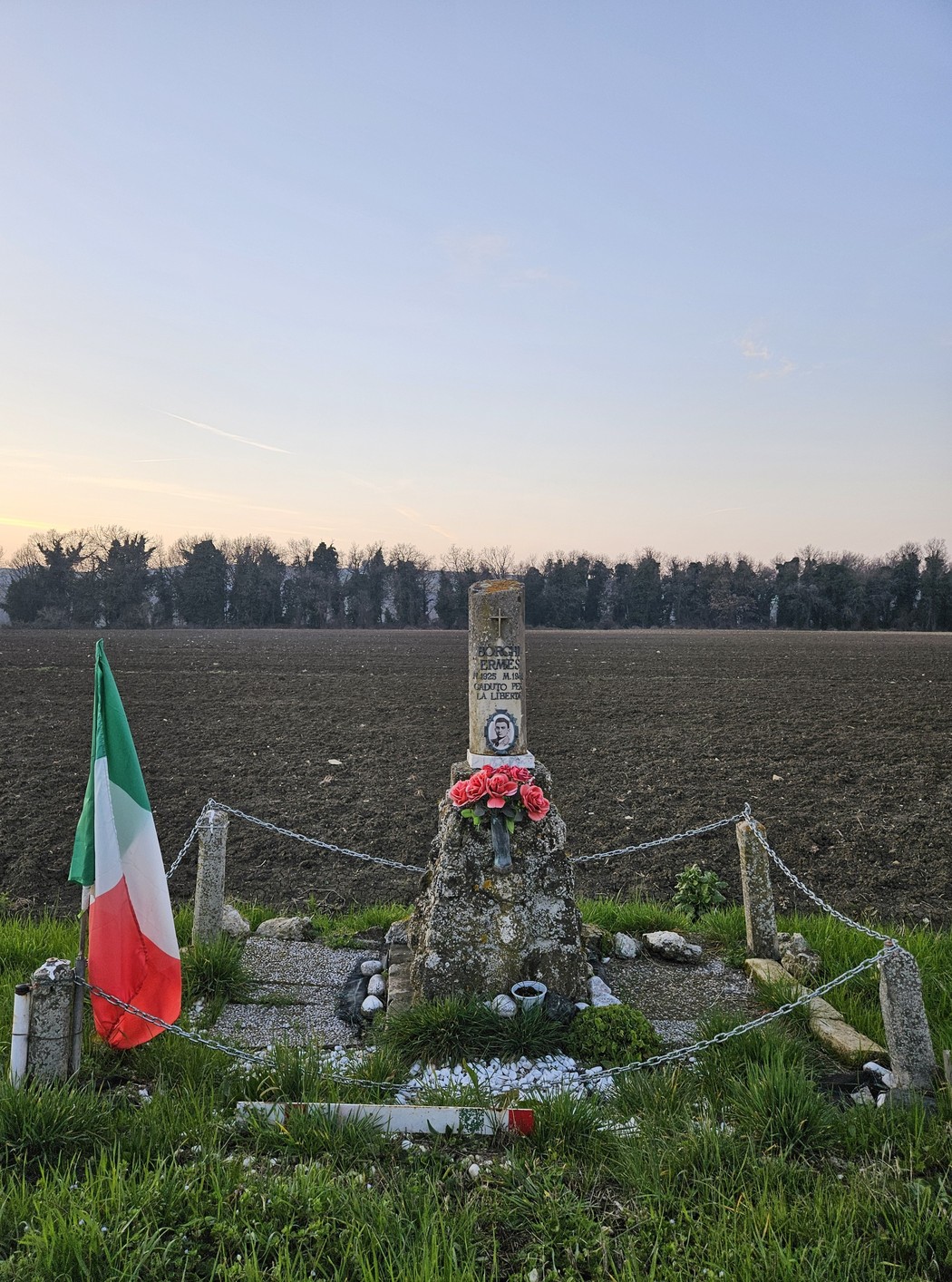 Cippo commemorativo con fiori, bandiera italiana e paesaggio rurale sullo sfondo.
