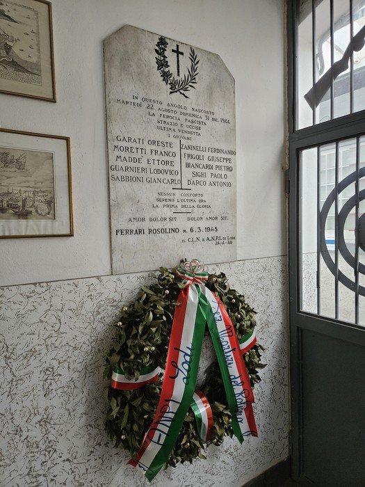 Lapide marmorea con iscrizione e croce, affissa a parete, con corona di alloro e nastri tricolore sottostante.