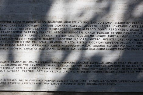 Monumento ai Caduti con iscrizioni commemorative su una superficie in pietra. Ombre delicate sovrastano il memoriale.