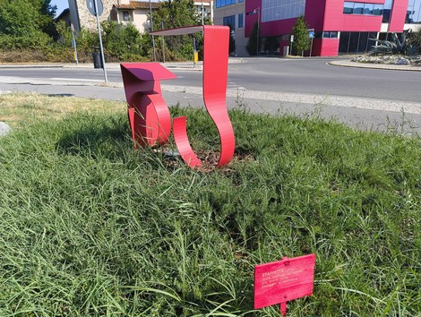 Scultura astratta rossa su prato verde, simbolo di resilienza femminile in un contesto urbano.