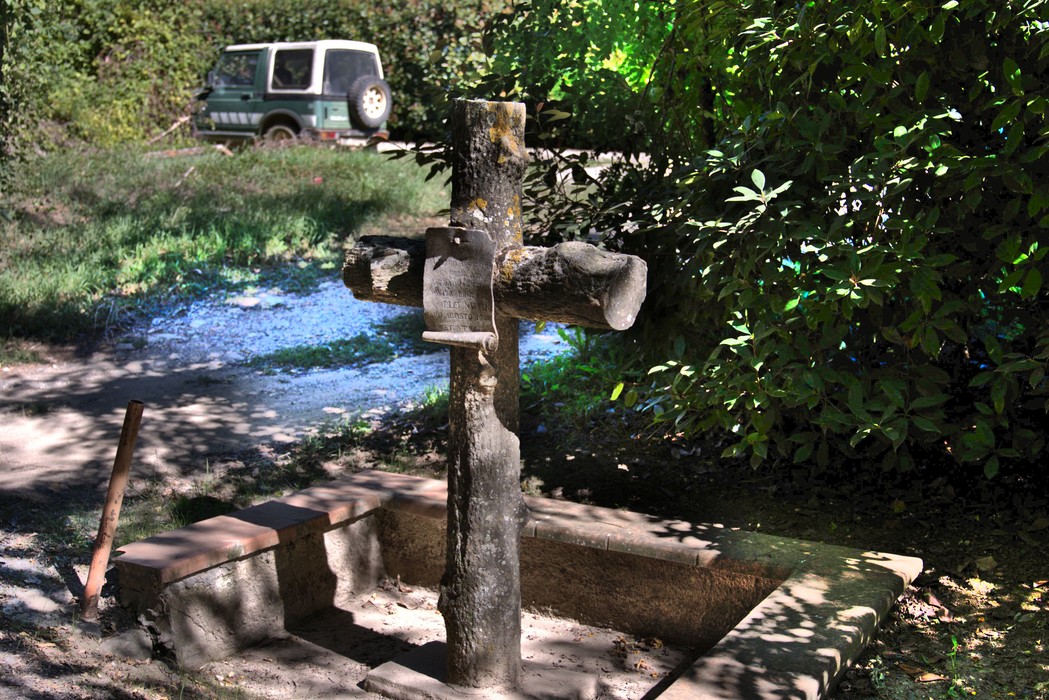 Croce in legno con base in pietra, in un'area erbosa immersa nel verde, con un fuoristrada sullo sfondo.