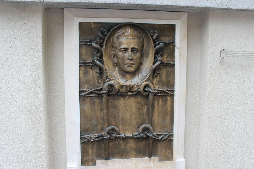 Monumento a D'Amico, bas-relief con volto maschile circondato da elementi simbolici di commemorazione.