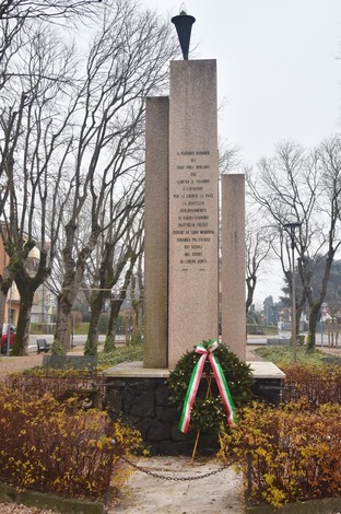 Monumento ai Caduti immerso nella vegetazione, con una corona d'alloro e bandiere tricolori. Atmosfera solenne.