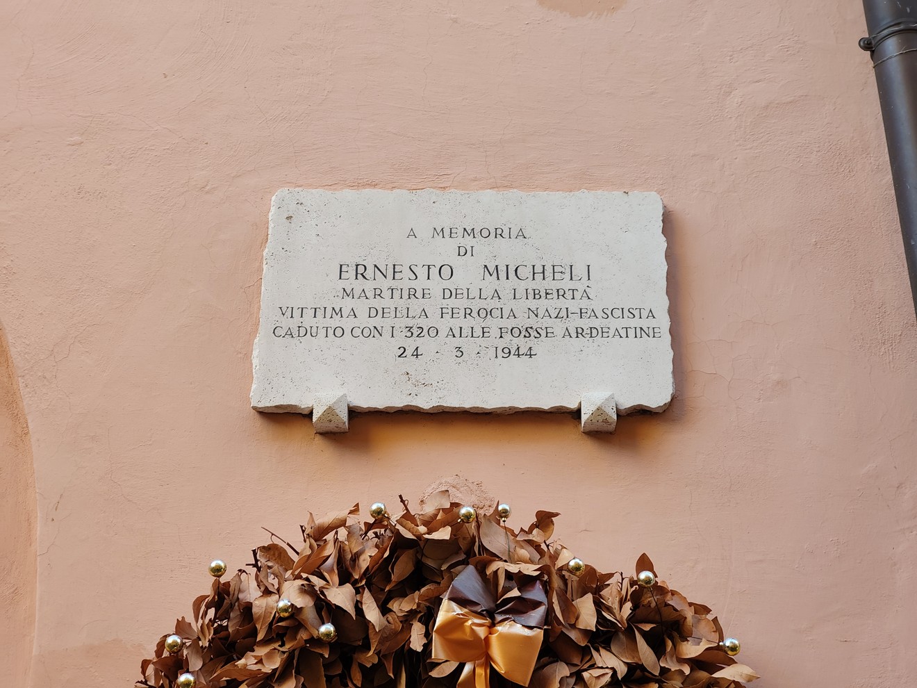 Lapide in pietra con iscrizione, fissata su una parete intonacata sopra una corona di fiori secchi.
