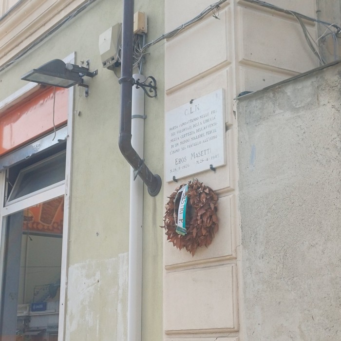 Lapide in marmo con iscrizione sulla facciata di un edificio, sormontata da una corona di foglie secche.
