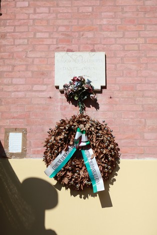 Lapide commemorativa a Orsini, con una corona di fiori e nastri decorativi, collocata su un muro di mattoni.