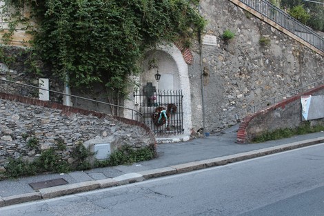 Edicola dei caduti di San Gottardo, affacciata su una strada, circondata da vegetazione e muretti in pietra.