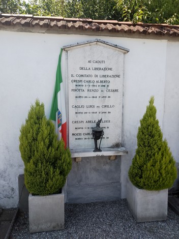 Lapide commemorativa ai caduti della Liberazione, con vegetazione e bandiera italiana in background.