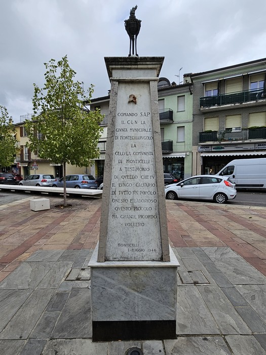 Monumento ai Partigiani, obelisco con decorazioni e iscrizioni, circondato da alberi e edifici.