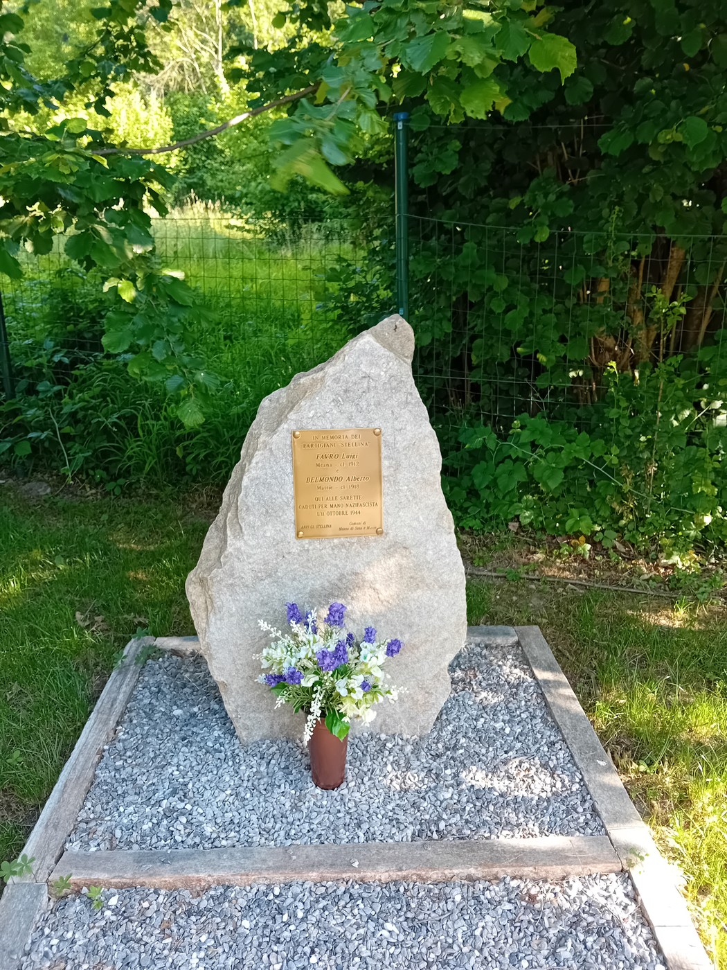 Cippo commemorativo con fiorellini, circondato da verde e pietre, in un'area tranquilla e naturale.