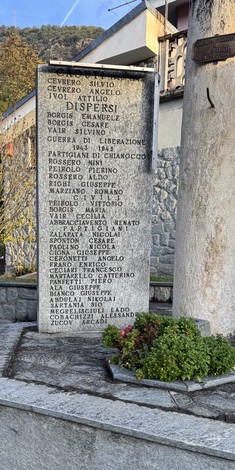 Stele in pietra con lunga iscrizione ai caduti, posta all'aperto davanti a un edificio, con cespuglio fiorito alla base.