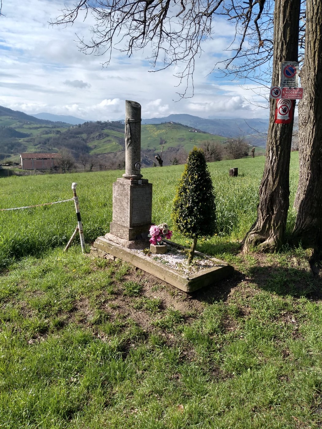 Cippo commemorativo immerso in un paesaggio collinare verdeggiante, con fiori e alberi circostanti.