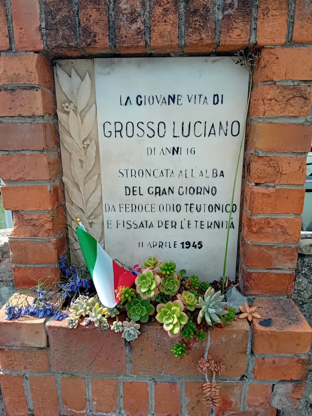 Lapide in mattoni commemorativa dedicata a Grosso Luciano, con fiori e bandiera italiana.