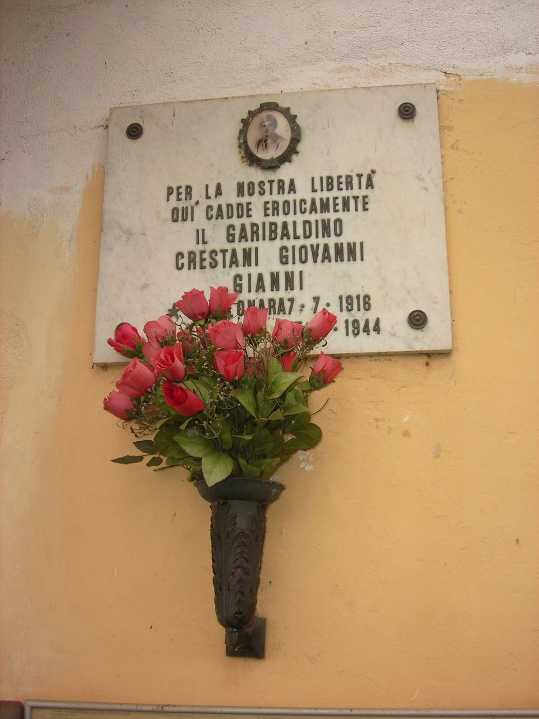 Lapide dedicata a Giovanni Crestani, garibaldino caduto per la libertà, con fiori rossi ornamentali.