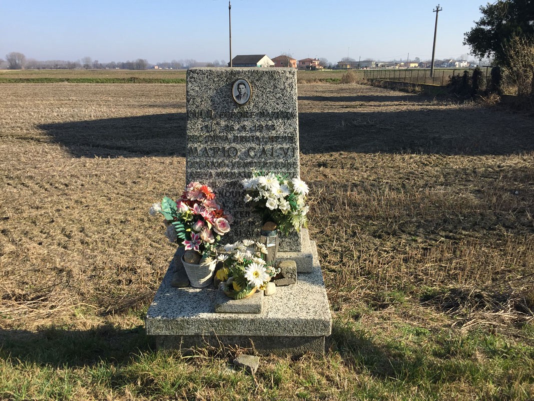 Cippo commemorativo a Calvi, circondato da fiori, in un paesaggio rurale.