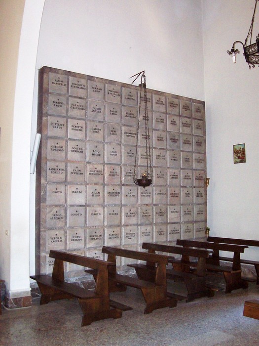 Cappella dei caduti con parete commemorativa e panche in legno per i visitatori. Atmosfera di riflessione e memoria.