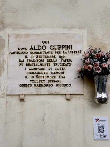 Lapide commemorativa dedicata ad Aldo Cuppini, partigiano, situata su una parete, circondata da fiori artificiali.