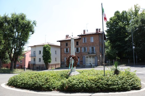 Monumento ai caduti in una rotonda, circondato da vegetazione e bandiera italiana, con edifici storici sullo sfondo.