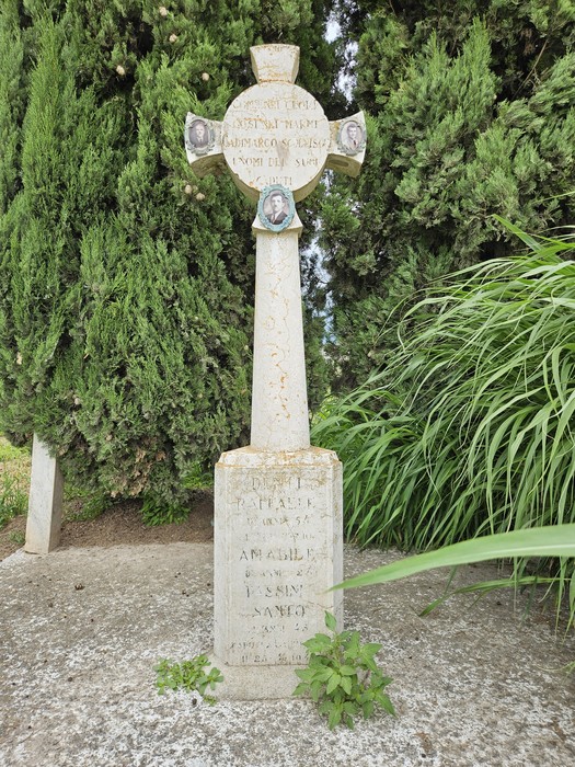 Cippo in pietra con croce, circondato da vegetazione, commemorativo di persone scomparse.