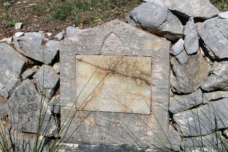 Cippo commemorativo a Perlamagna, circondato da pietre e vegetazione.