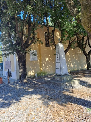 Lapide commemorativa in un giardino, con alberi verdi e una parete di fondo gialla. Atmosfera tranquilla e storica.