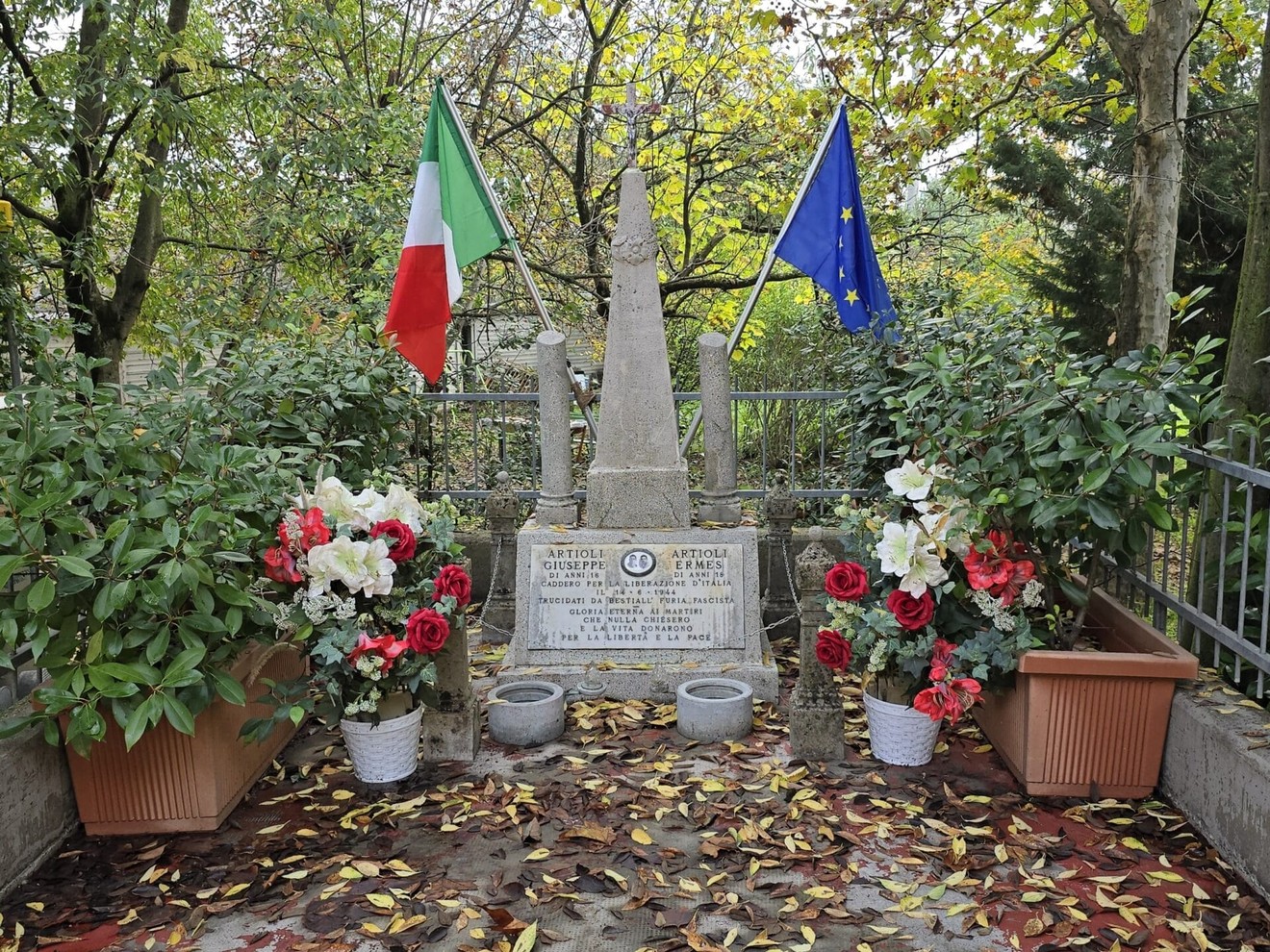Cippo commemorativo con fiori e bandiere italiane ed europee, immerso in un contesto naturale.
