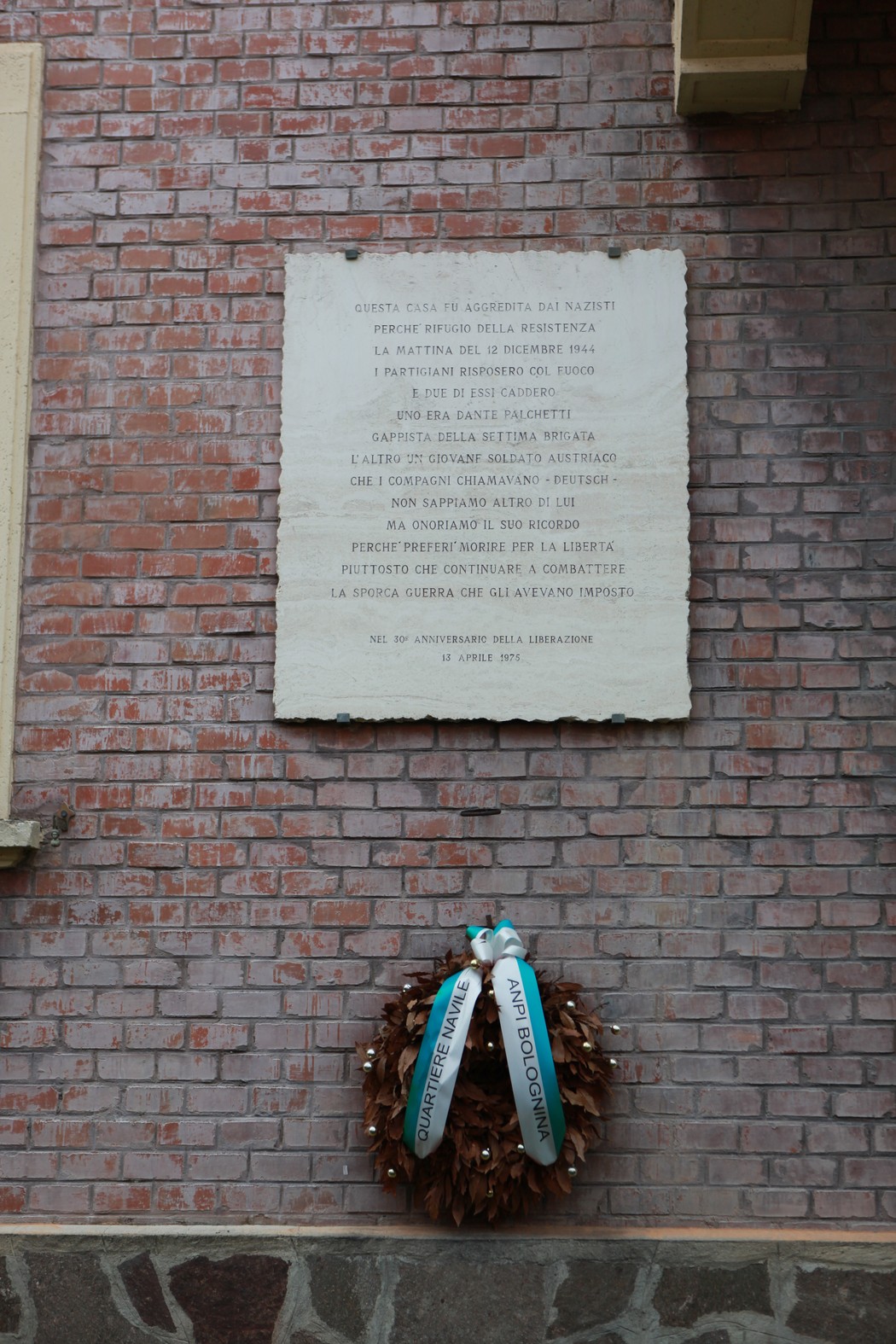 Lapide commemorativa di una base partigiana con una corona di alloro ai piedi, affissa su un muro di mattoni.