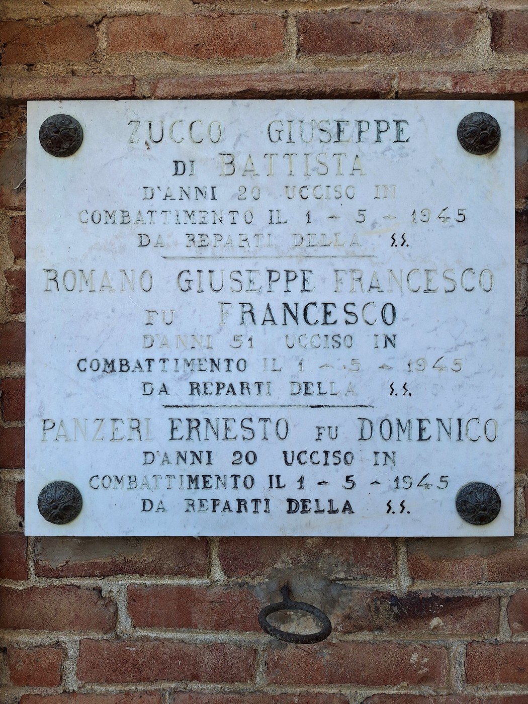 Lapide marmorea con iscrizione, fissata a una parete in mattoni con quattro rosette angolari.