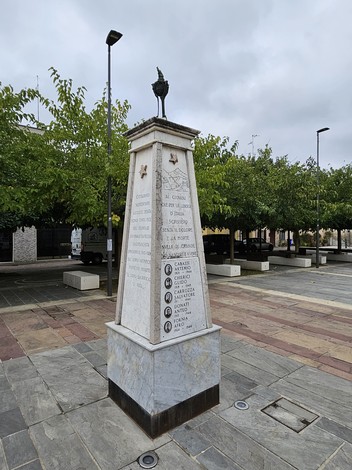 Monumento ai Partigiani, con obelisco di marmo e simboli, circondato da alberi e strade cittadine.