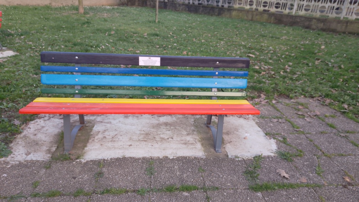 Panchina colorata nel parco Donne della Resistenza, circostante verde e ambiente tranquillo.