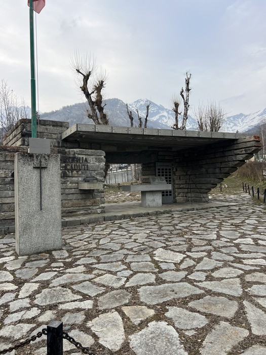 Monumento in pietra e cemento con tettoia sporgente, pilastro con croce inciso, su piazzale lastricato tra montagne.