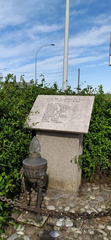 Stele commemorativa circondata da vegetazione, con lampada e dedicata ai caduti di Le Budrie. cielo sereno sullo sfondo.