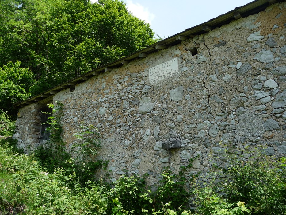 Lapide storica sulla parete di una cascina in pietra, circondata da vegetazione lussureggiante.