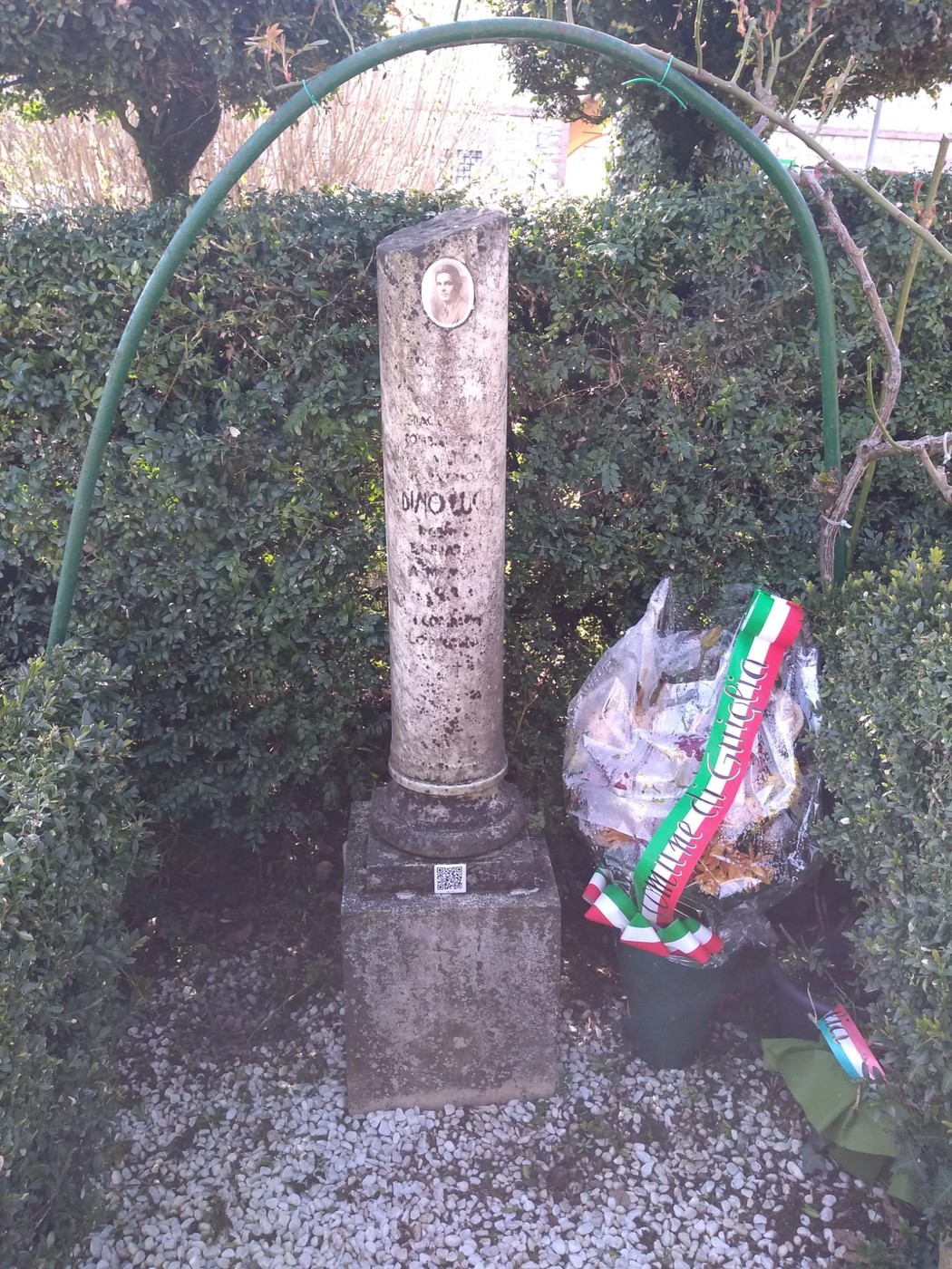Cippo commemorativo in un giardino, con fiori e decorazioni, circondato da siepi verdi.