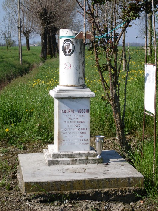 Cippo commemorativo dedicato a Gisberto Vecchi, situato in un'area rurale con vegetazione circostante.
