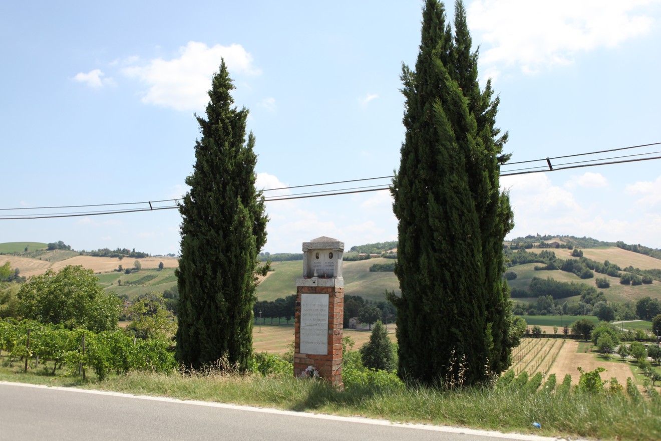 Cippo commemorativo tra due cipressi, affacciato su un paesaggio collinare e agricolo.