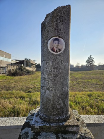 Cippo commemorativo di Guatelli con foto e iscrizioni, circondato da un'area verde e un cielo sereno.