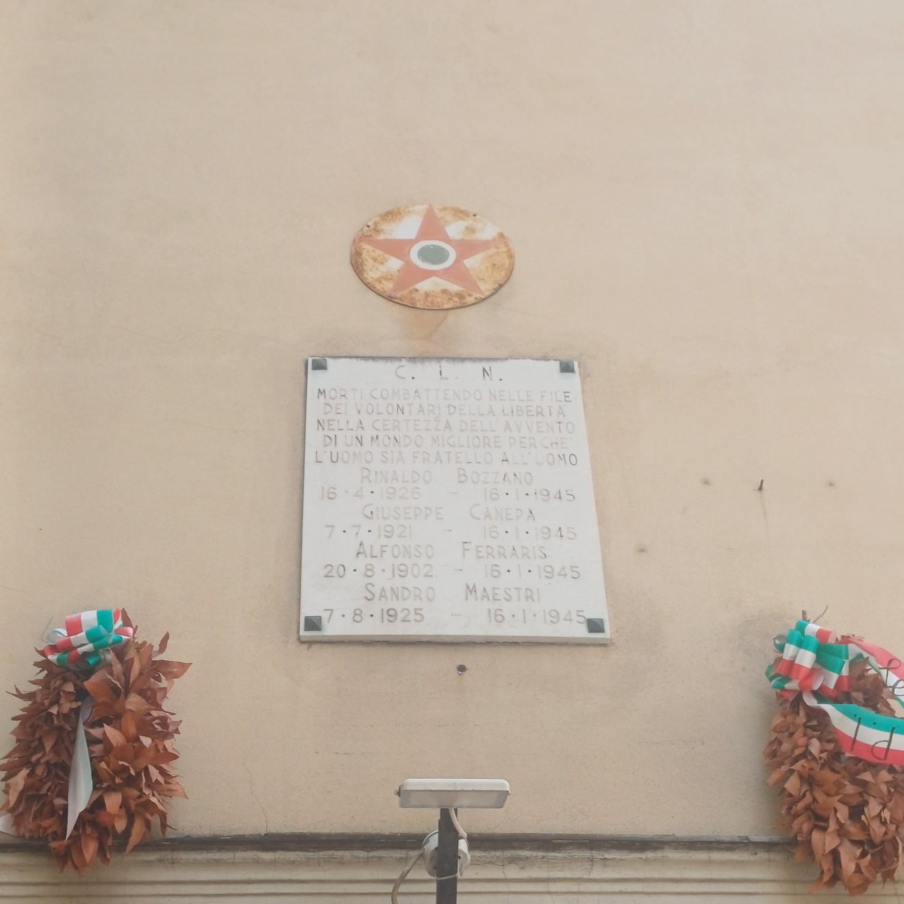 Lapide marmorea con iscrizione su parete, sormontata da una stella decorativa, affiancata da due corone tricolori.
