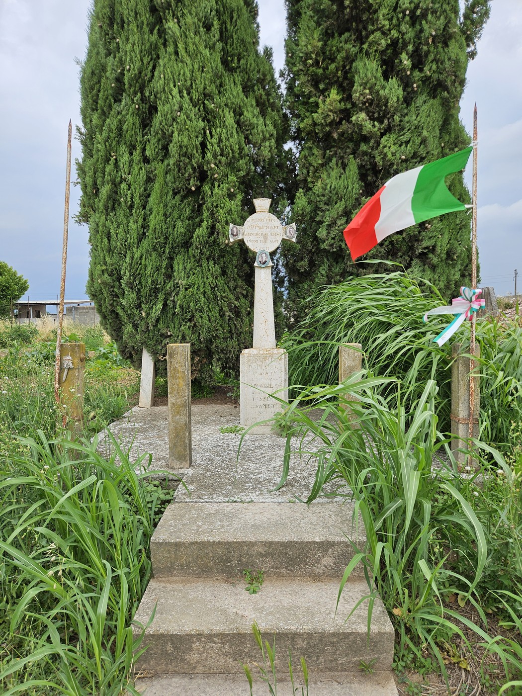 Cippo commemorativo circondato da cespugli e alberi, con bandiera italiana che sventola.