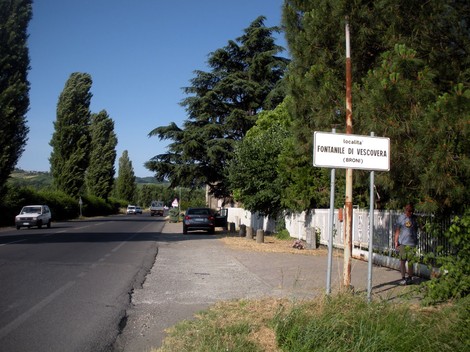 Cippo commemorativo in un contesto stradale, circondato da alberi e una recinzione. Atmosfera di memoria storica.