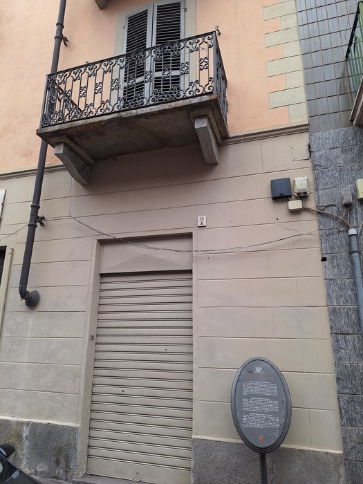 Facciata di edificio con balcone in ferro battuto, saracinesca al piano terra e targa ovale accanto all'ingresso.