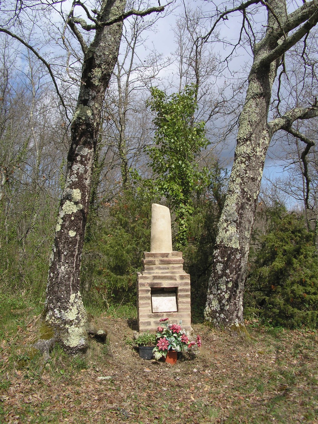 Cippo commemorativo incastonato tra due alberi, circondato da fiori in un ambiente naturale tranquillo.