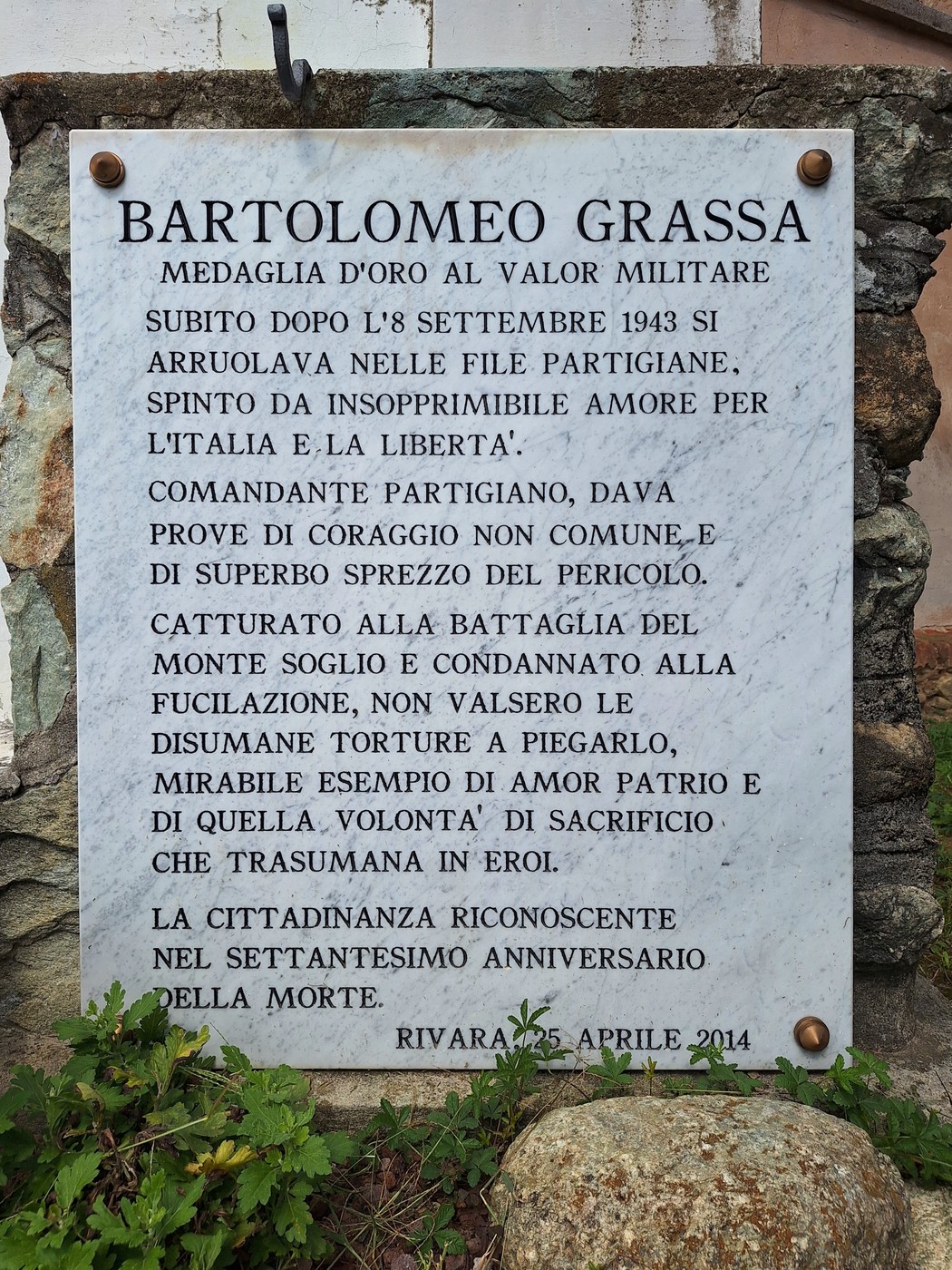 Lapide commemorativa in marmo con iscrizione, fissata a un muro di pietra e circondata dalla vegetazione.