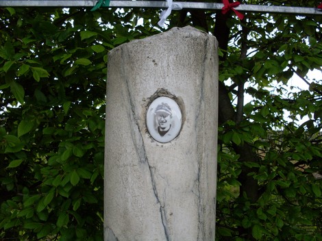 Cippo in pietra con un medaglione, circondato da vegetazione. Monumento evocativo della memoria storica.
