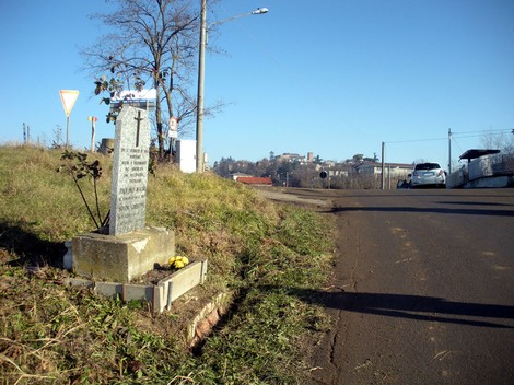 Cippo commemorativo situato lungo una strada, circondato da erba e con vista su un paesaggio urbano.
