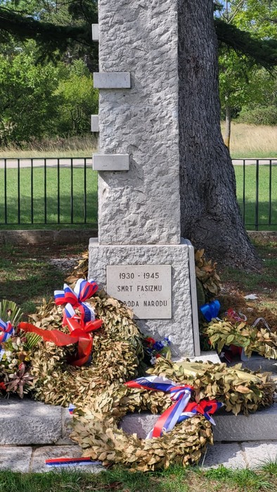 Cippo in pietra con lapide incisa, ai piedi un albero e corone di alloro con nastri tricolore.