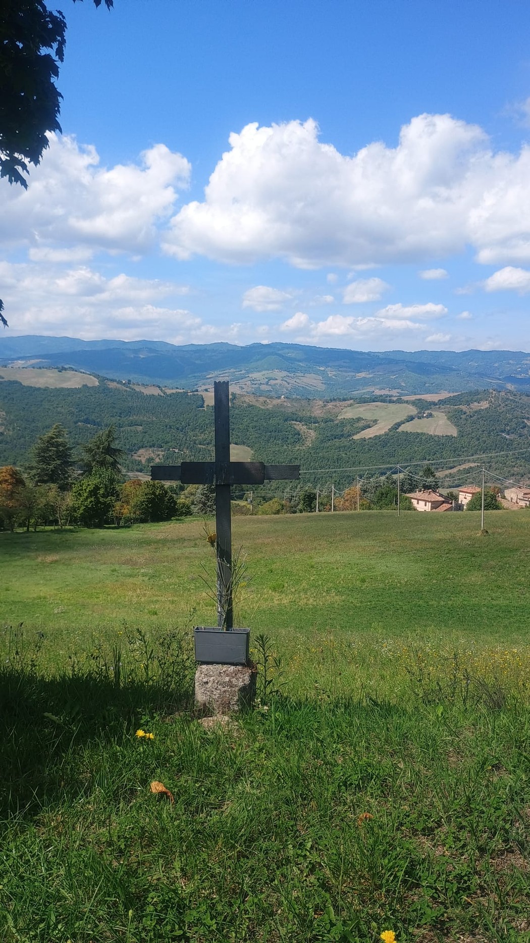 Vista panoramica con croce in legno su un prato verde, circondata da colline e un cielo blu con nuvole bianche.